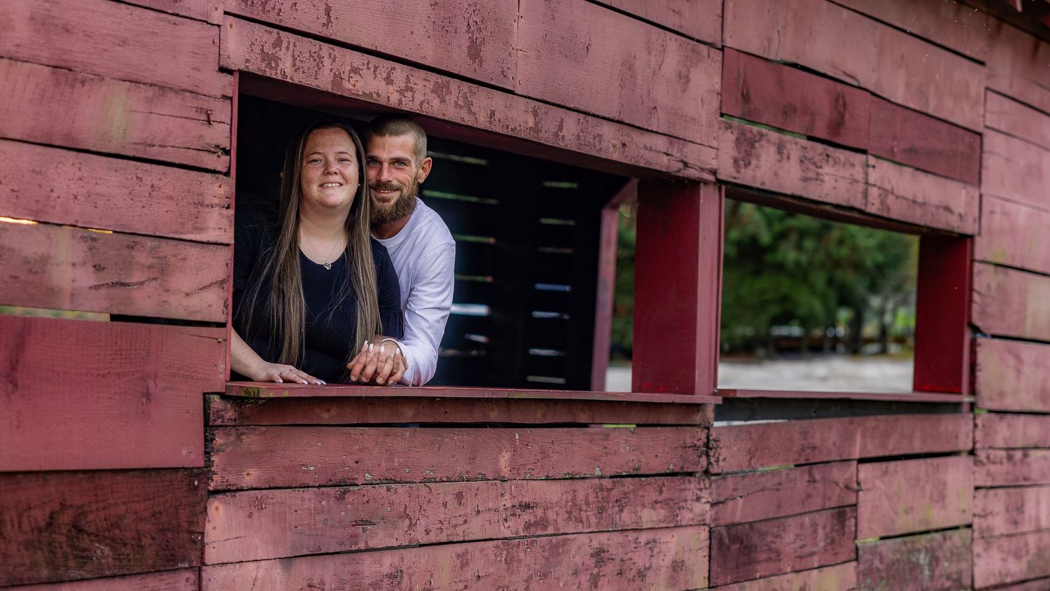 A rustic red barn with windows serves as a backdrop for a romantic engagement photo session.