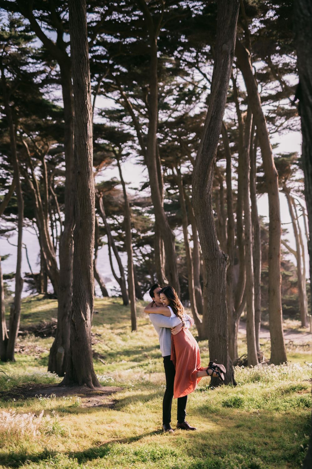 Couple embracing romantically in a sunlit forest with tall trees and green grass.