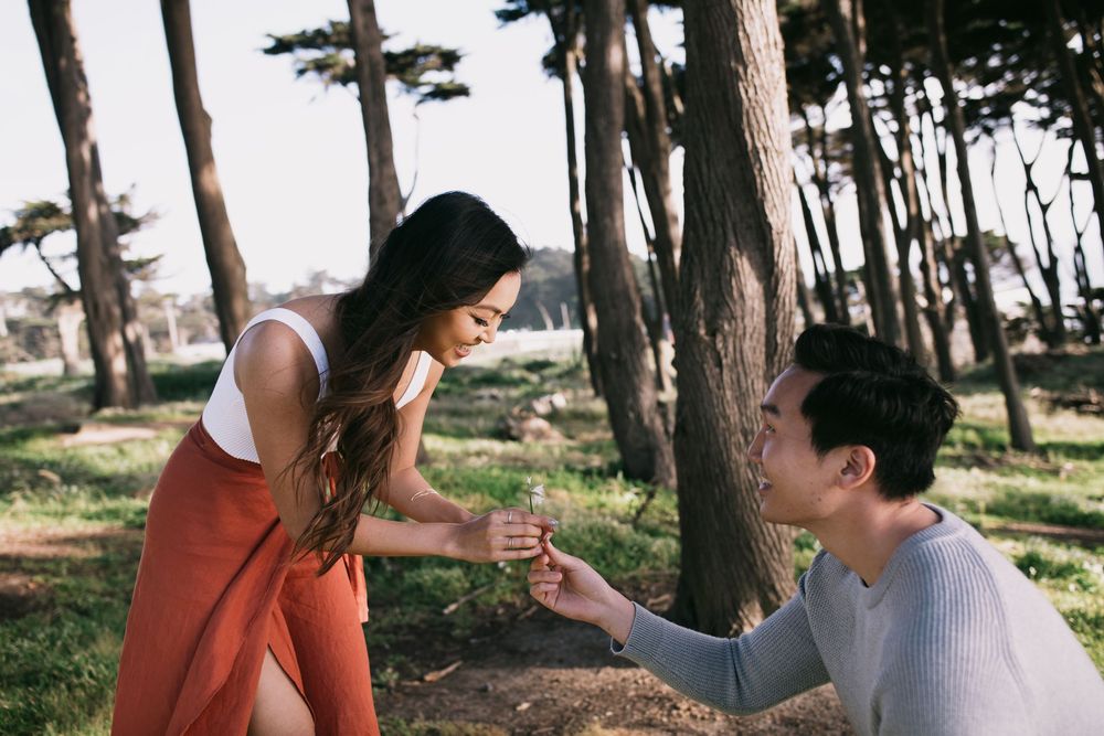 Man kneeling on one knee proposing to woman outdoors among trees in a romantic marriage proposal scene.