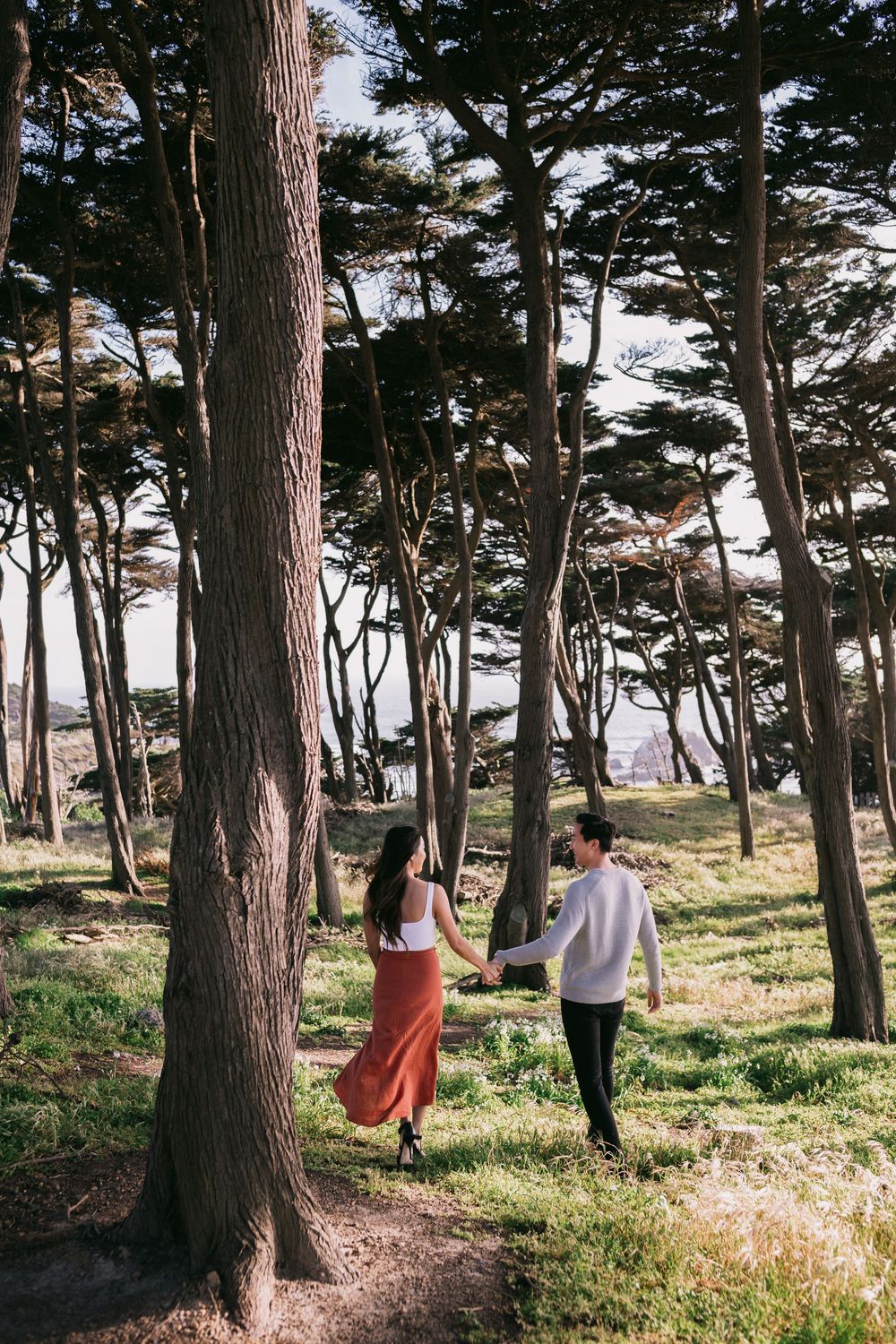 Couple holding hands walking through a sunlit cypress forest near the San Francisco coast.