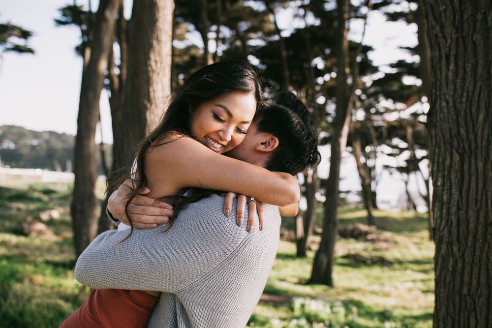 A couple shares a warm embrace outdoors among tall trees, smiling joyfully in a sunlit forest setting.