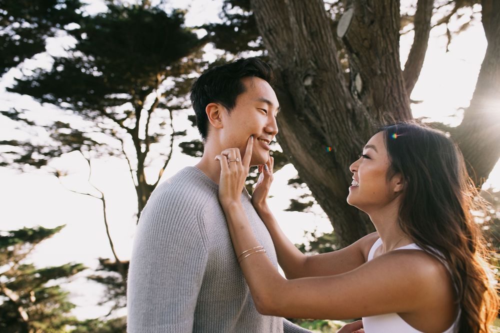 Happy Asian couple laughing and touching faces outdoors near trees in warm golden sunlight.