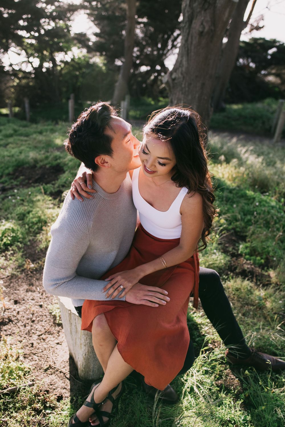 Romantic couple sitting on a stump in a forest, man kissing woman's cheek in golden light.