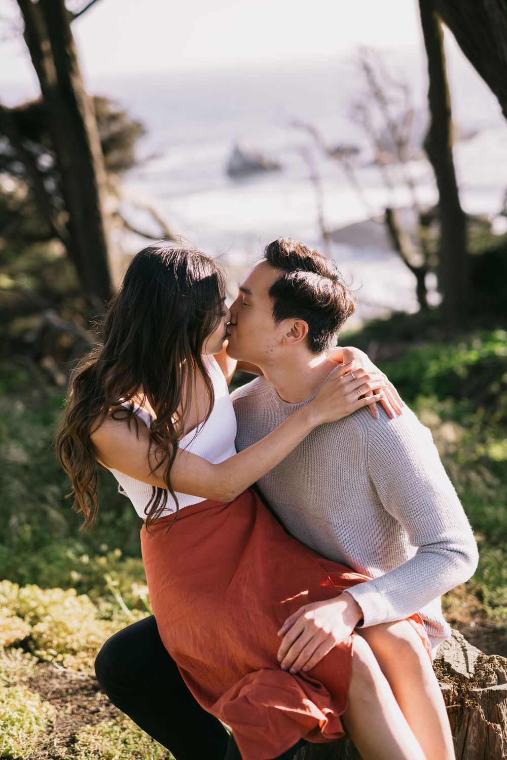 Couple sharing a tender kiss outdoors with a scenic coastal backdrop and cypress trees.