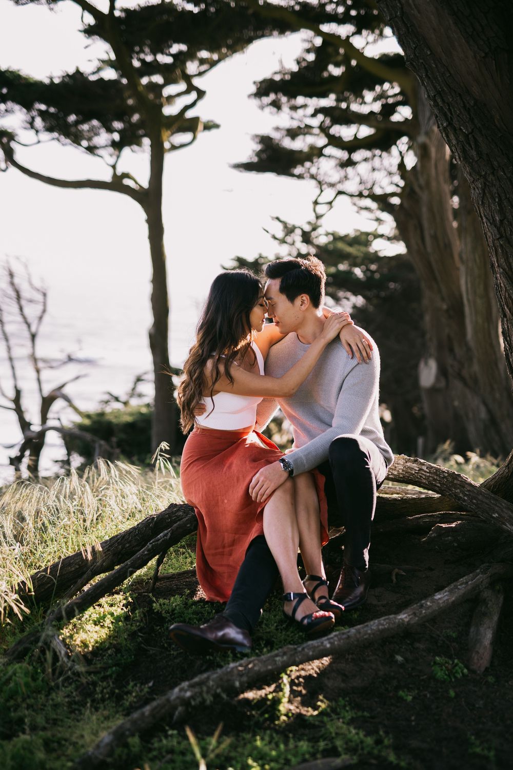A couple shares a romantic moment sitting on fallen tree branches surrounded by cypress trees near a coastal cliffside.