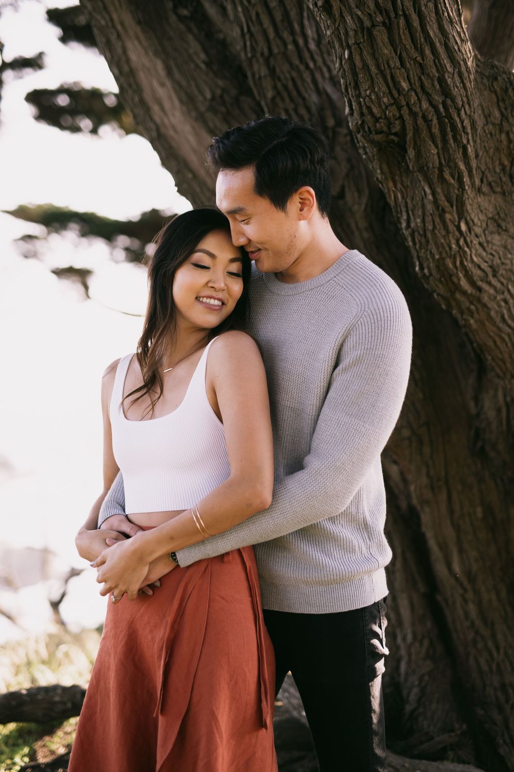 A couple embraces tenderly near a tree, woman in white top and rust skirt smiling, man in gray sweater holding her close.