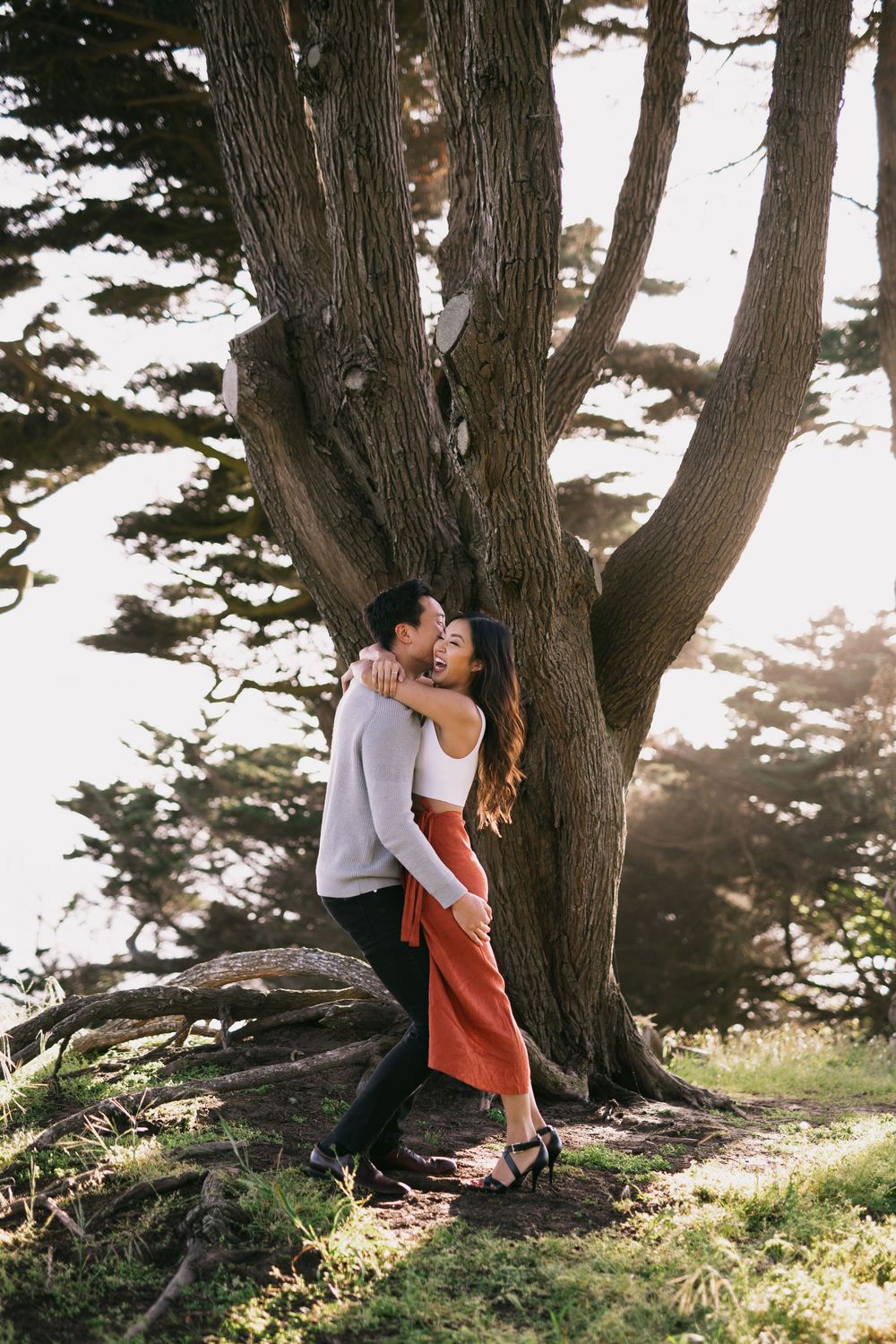 Couple laughing and hugging against a massive cypress tree in warm sunset light outdoors.