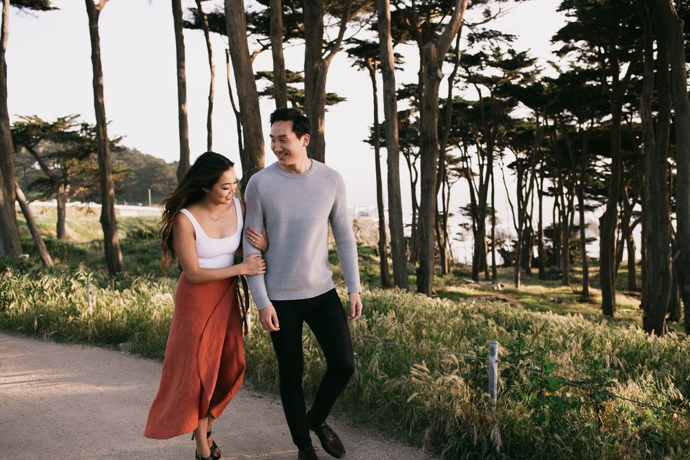 A couple walks hand in hand along a sunny path surrounded by tall trees and lush greenery.