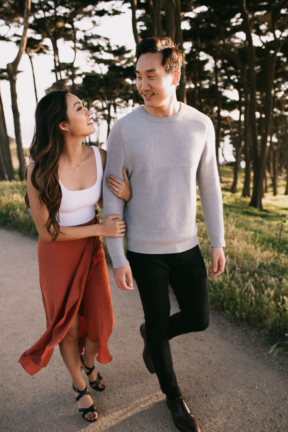 Couple walking arm in arm on a tree-lined path, smiling at each other in warm golden light.
