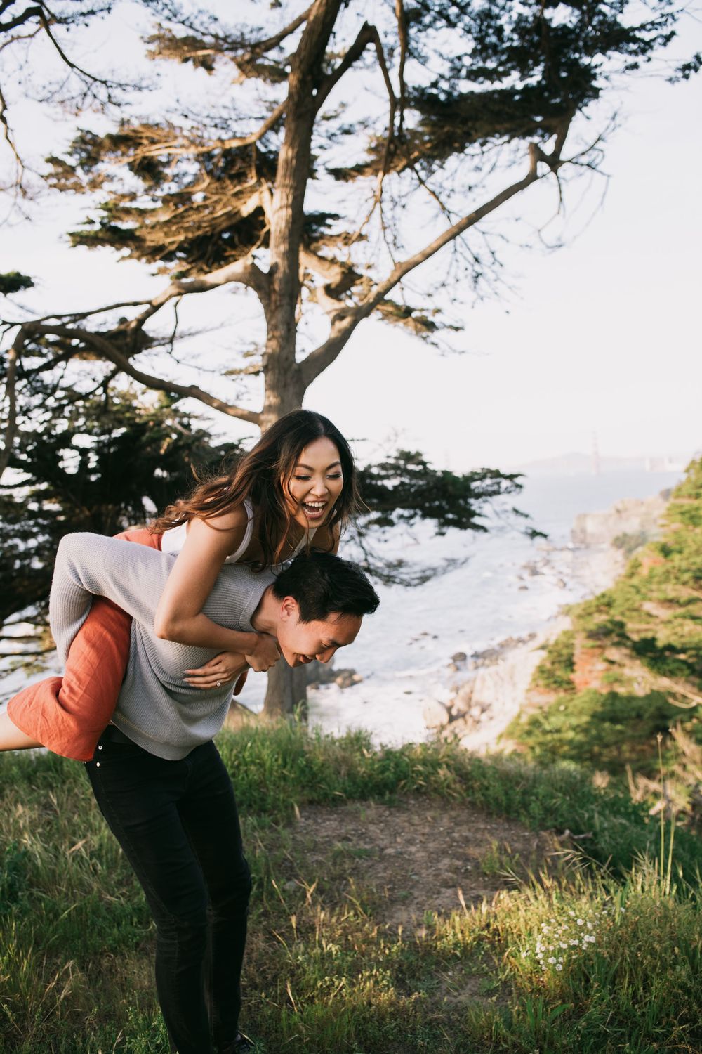 A joyful couple shares a piggyback ride on a grassy coastal cliff, laughing together beneath a tall tree.