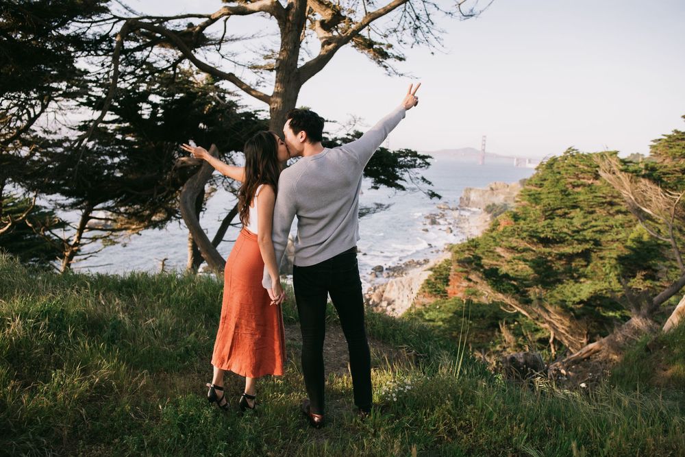 Couple pointing at the Golden Gate Bridge from a scenic overlook surrounded by trees and greenery.