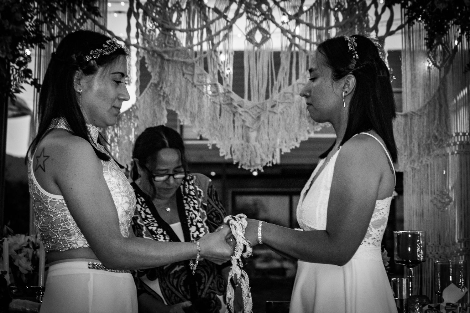Pareja de novias en ceremonia de matrimonio estilo boho detalle de unión de manos con cuerdas frente a un altar de macramé tejido
