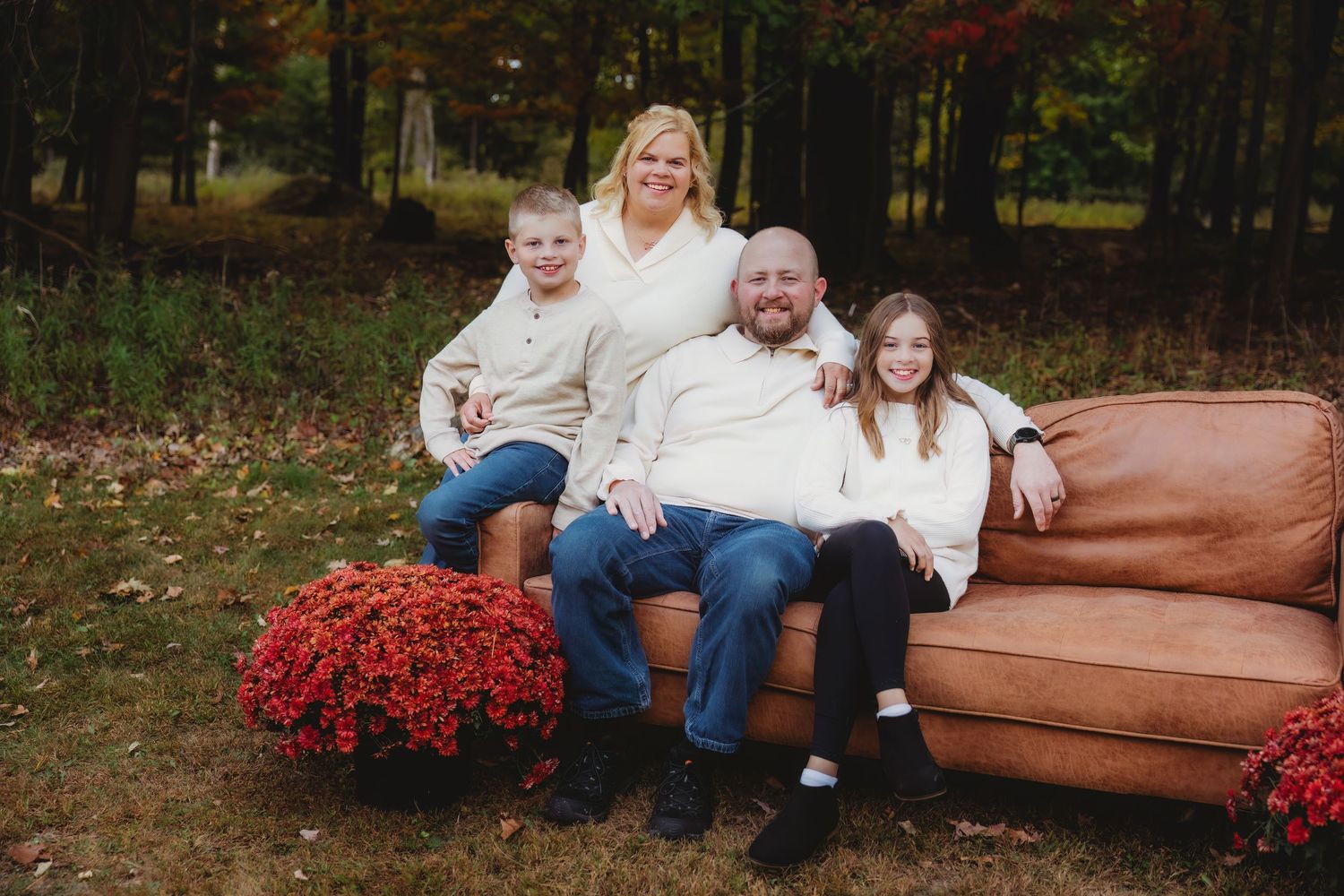 A family wearing coordinated cream and denim outfits sits together on fallen autumn leaves during an outdoor photoshoot.