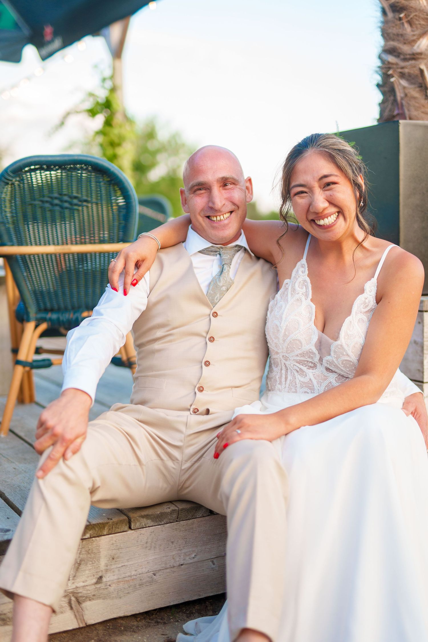 A couple in wedding attire smiles together while sitting outdoors at a venue with rattan furniture.