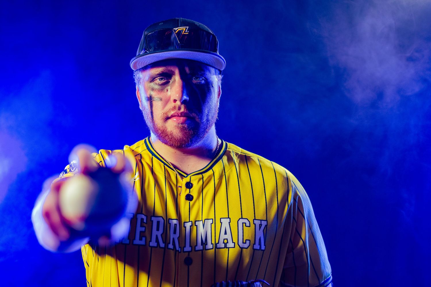 Merrimack baseball player in yellow pinstripe jersey pointing forward dramatically in blue smoky atmosphere.