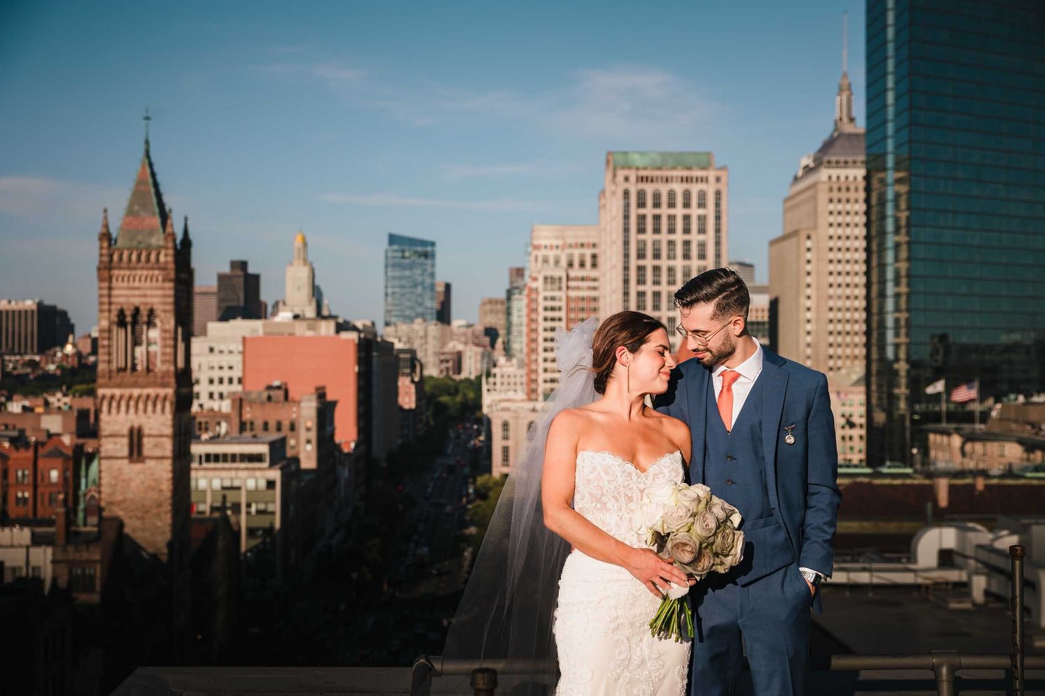 The bride and groom embrace for a romantic portrait atop the Lenox Hotel rooftop deck with the Boston Skyline behind them