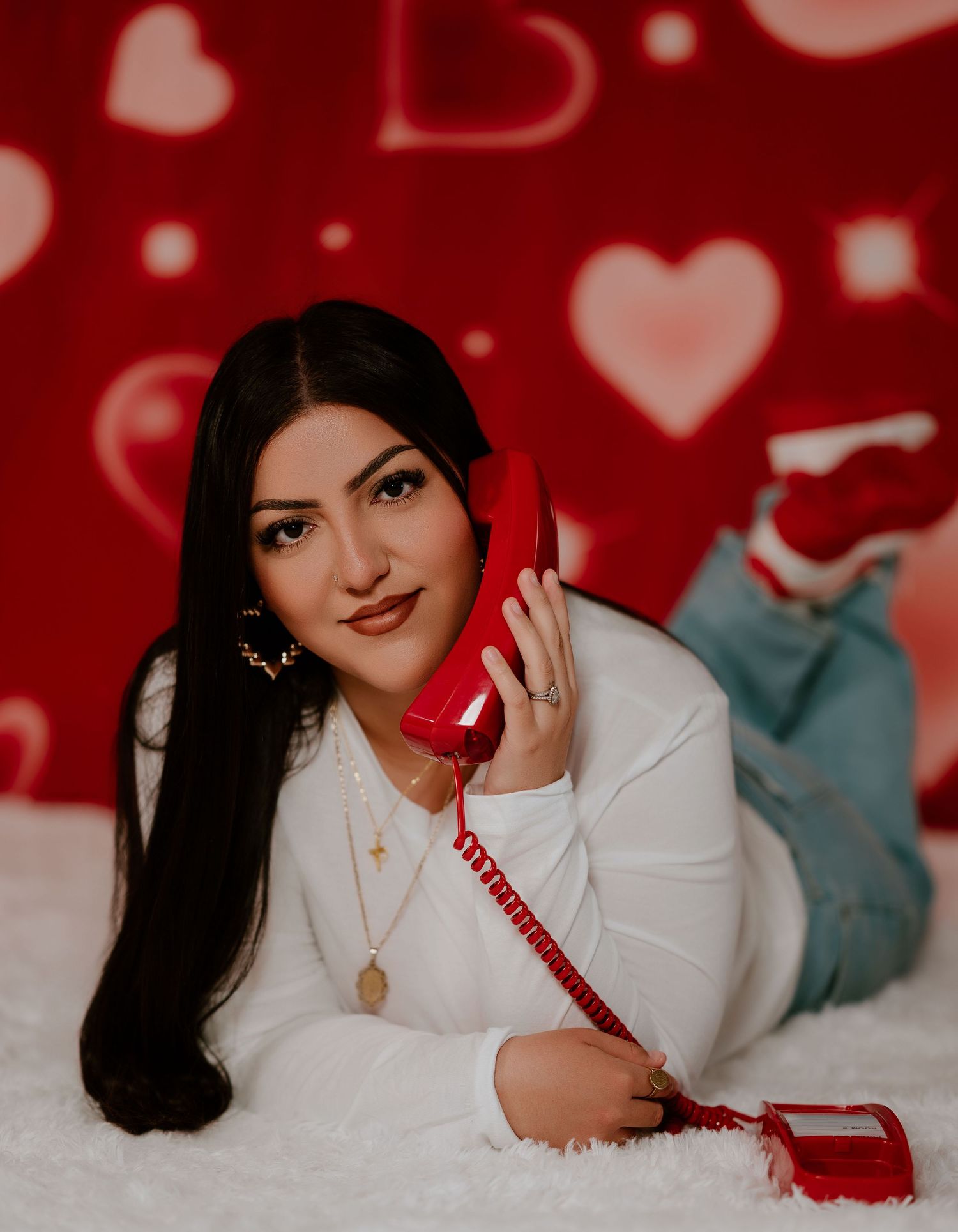 Person in white blazer lying on red background with vintage corded telephone and romantic heart decorations.