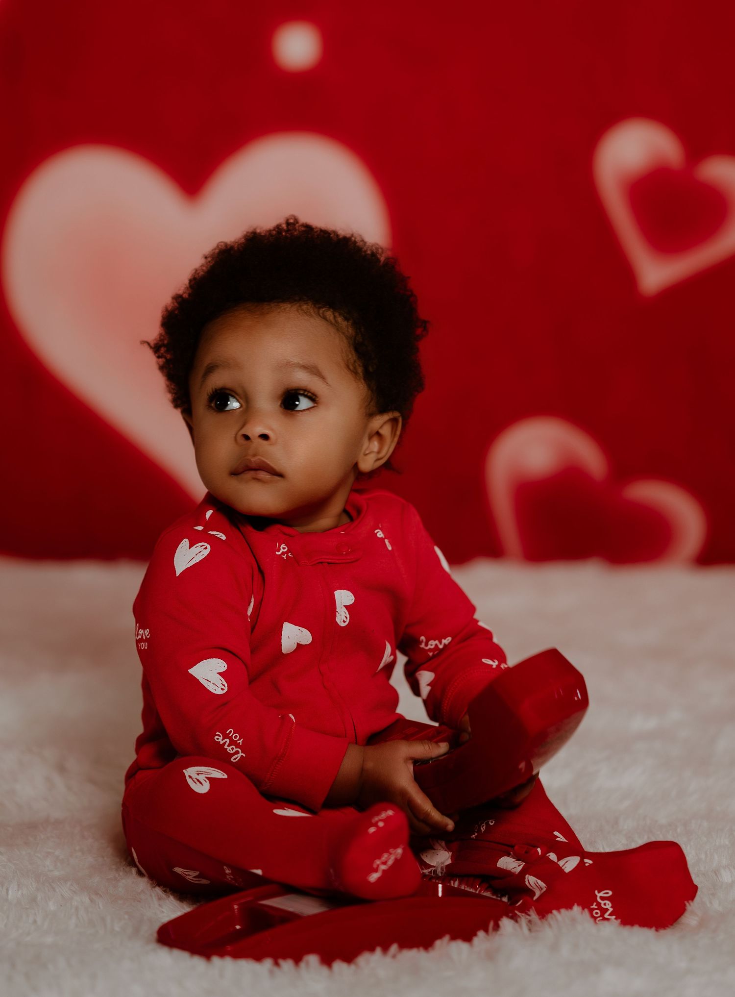 Baby in red heart-patterned outfit lies on white carpet with Valentine's Day themed backdrop.