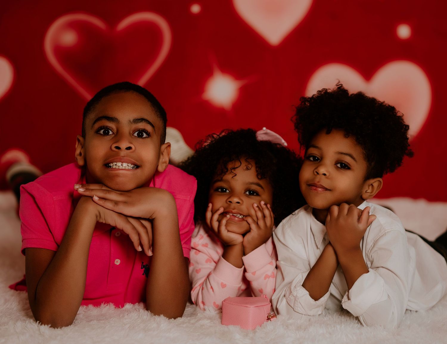 Three children pose together on white fur against a red background with floating heart shapes.