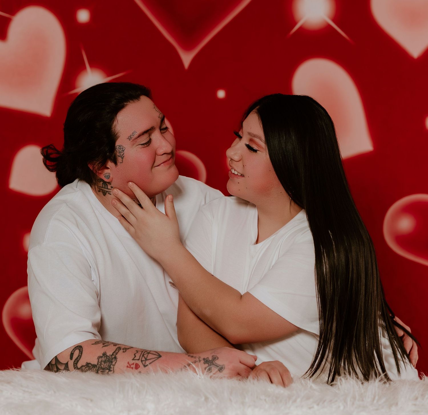 Romantic couple in white outfits embracing against red background with floating heart shapes during Valentine's photoshoot.