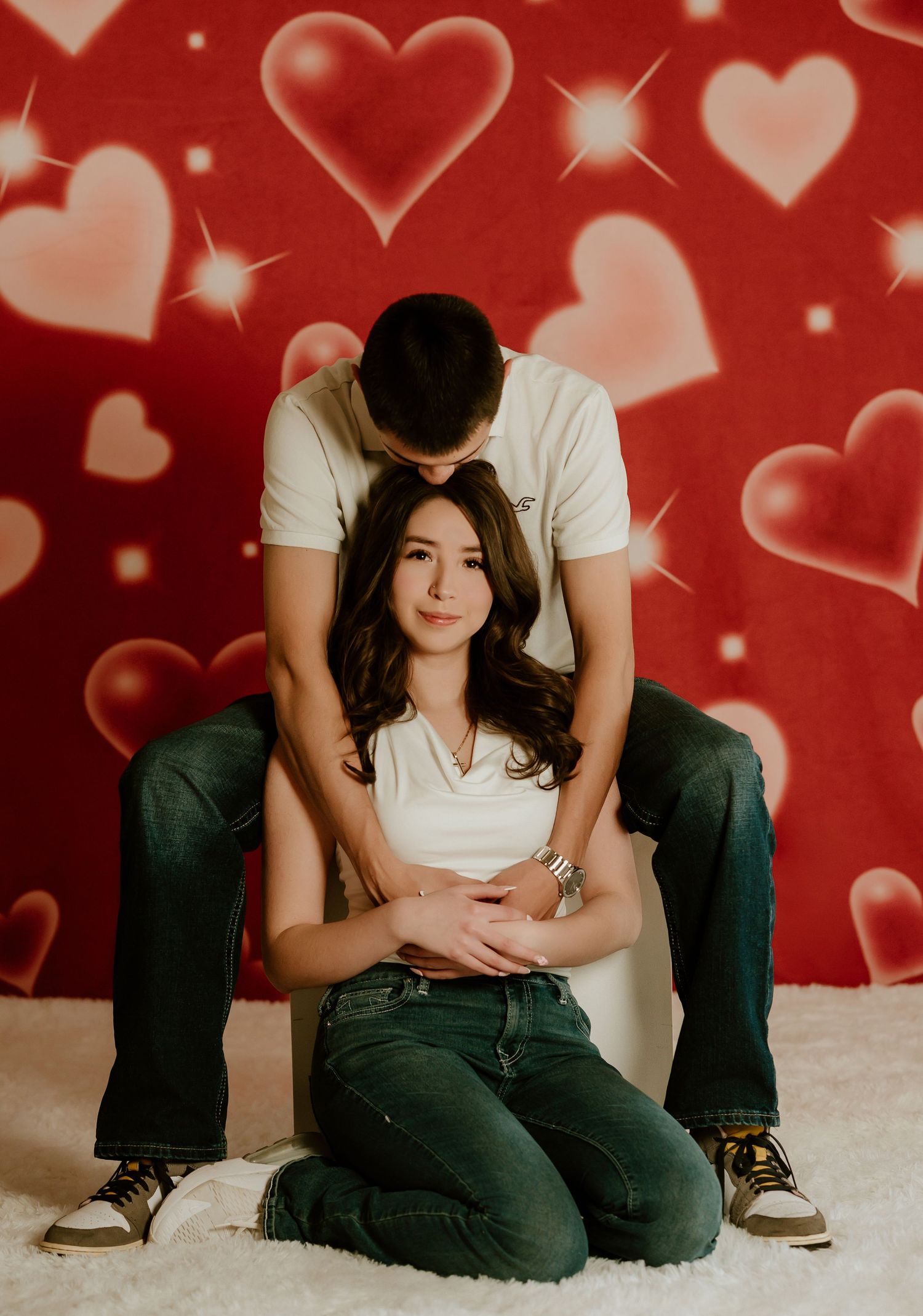 A romantic couple sits together against a red backdrop with floating heart designs and sparkles creating a Valentine's mood.