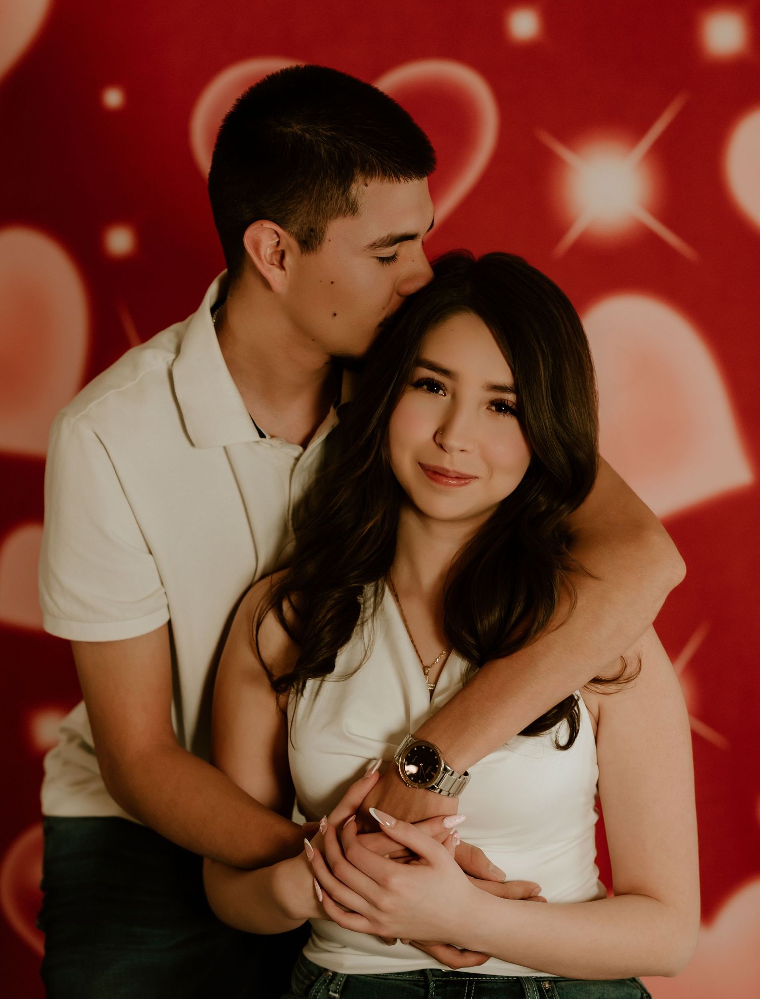 Romantic couple embraces against red background with heart bokeh and sparkle effects in Valentine's themed photo shoot.