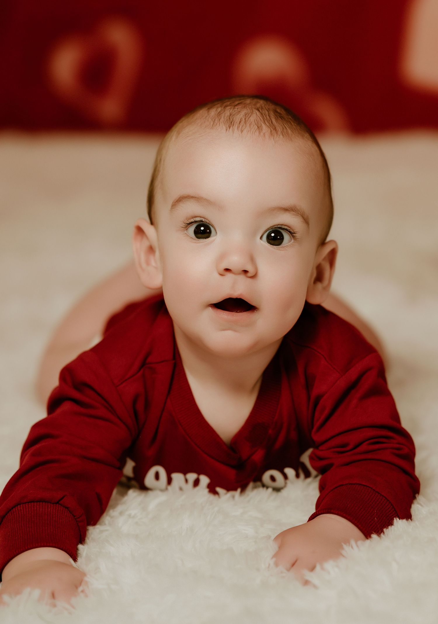 Baby in burgundy sweater lying on fluffy white carpet looking up with curious expression.