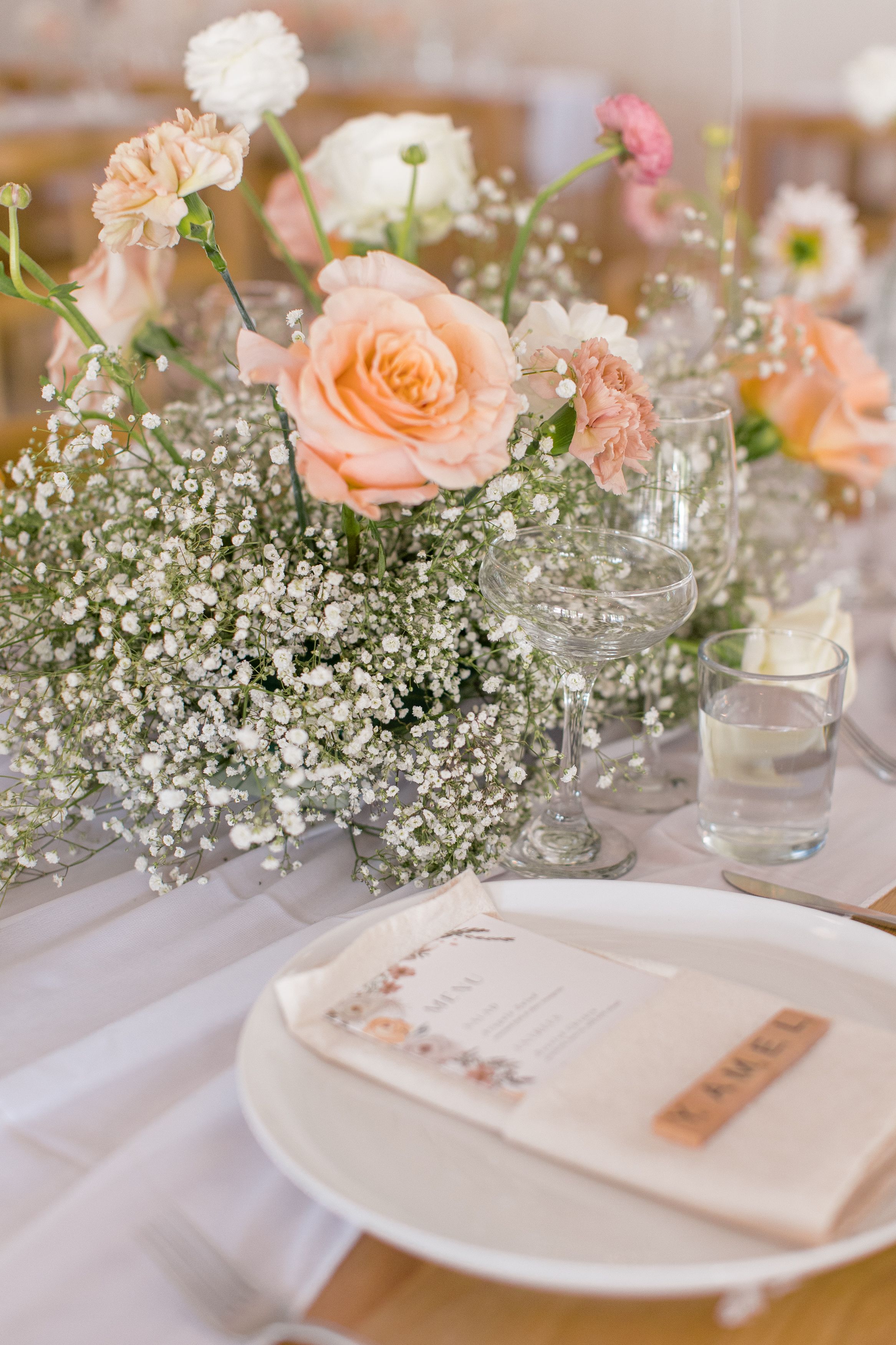 Elegant wedding reception place settings decorated with peach roses and baby's breath in crystal vases on a white tablecloth.