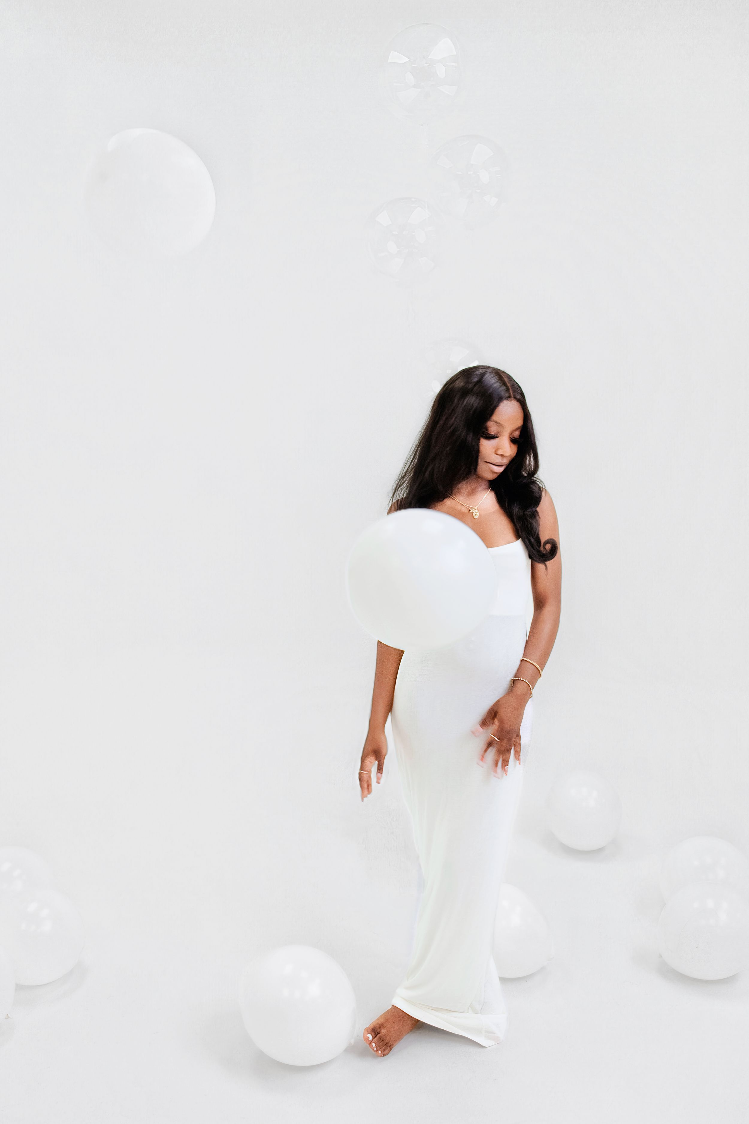 Person in flowing white dress posed gracefully with white balloon on light background in elegant photoshoot.