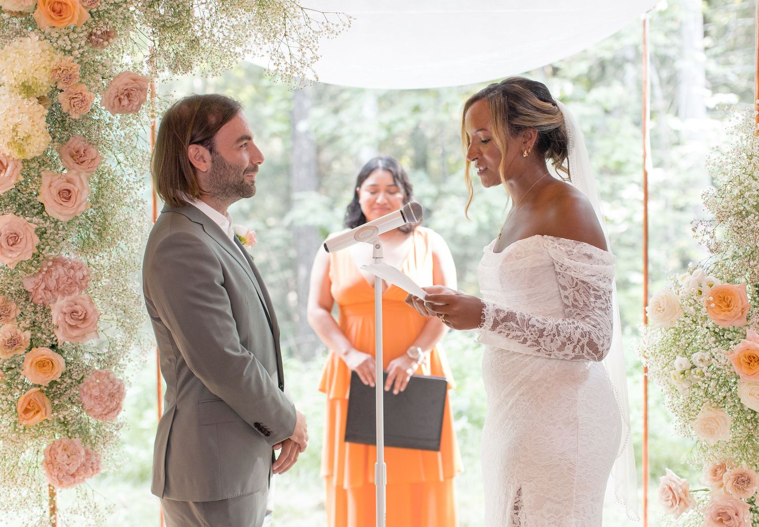 Wedding couple at rustic outdoor venue with peach and coral floral arch shares intimate moments during ceremony.