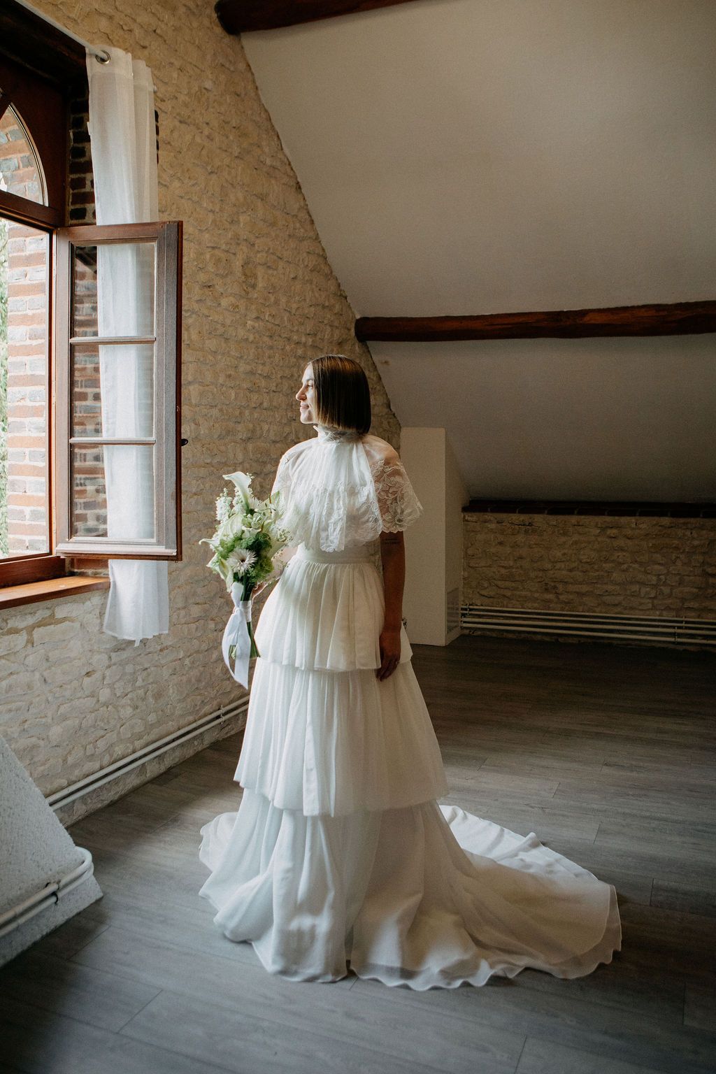Bride in tiered white lace wedding dress holding bouquet while gazing out arched window.