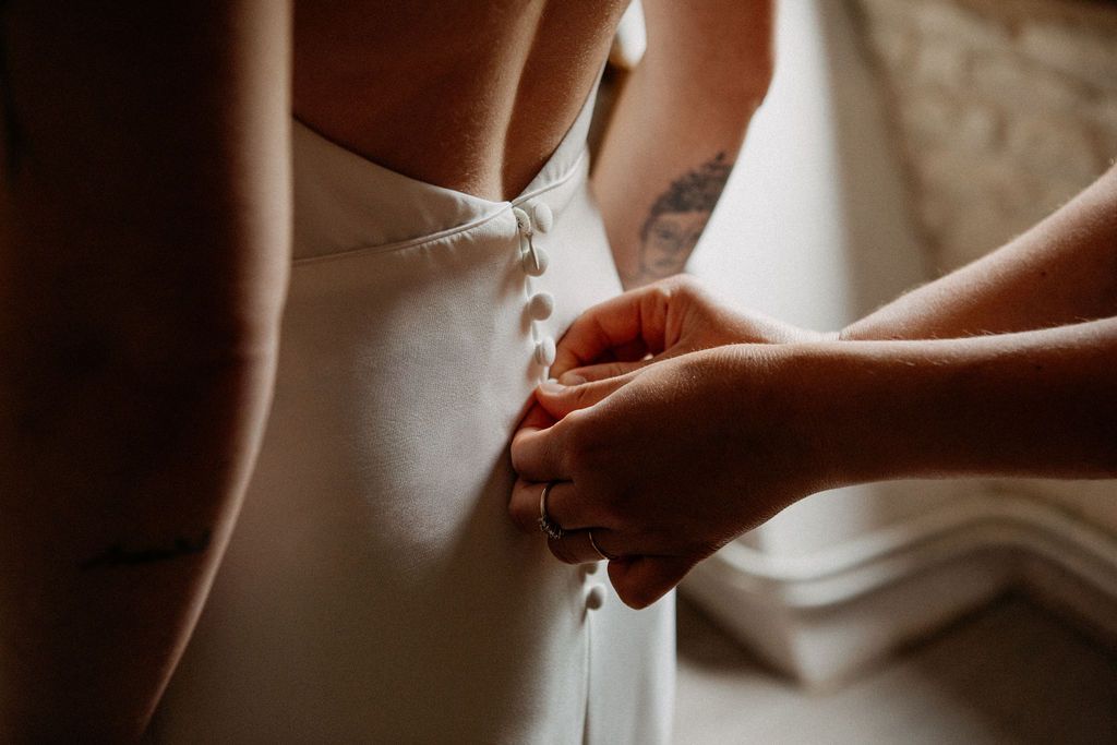 Close-up of tattooed hands fastening delicate buttons on back of white wedding dress.