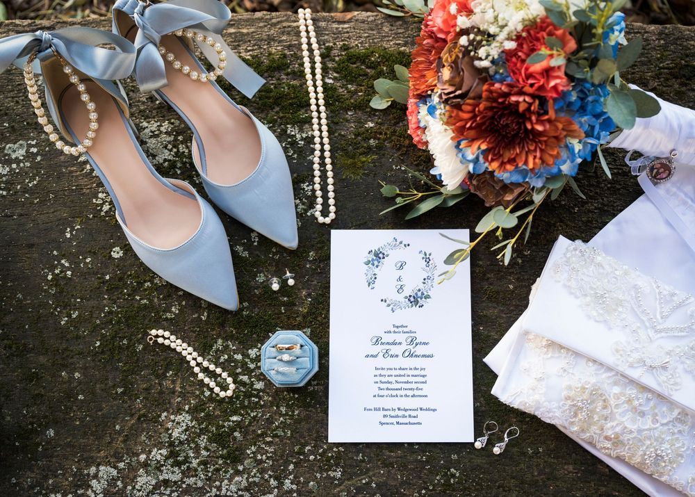 Wedding details including blue shoes, invitation, and floral elements arranged on stone surface.