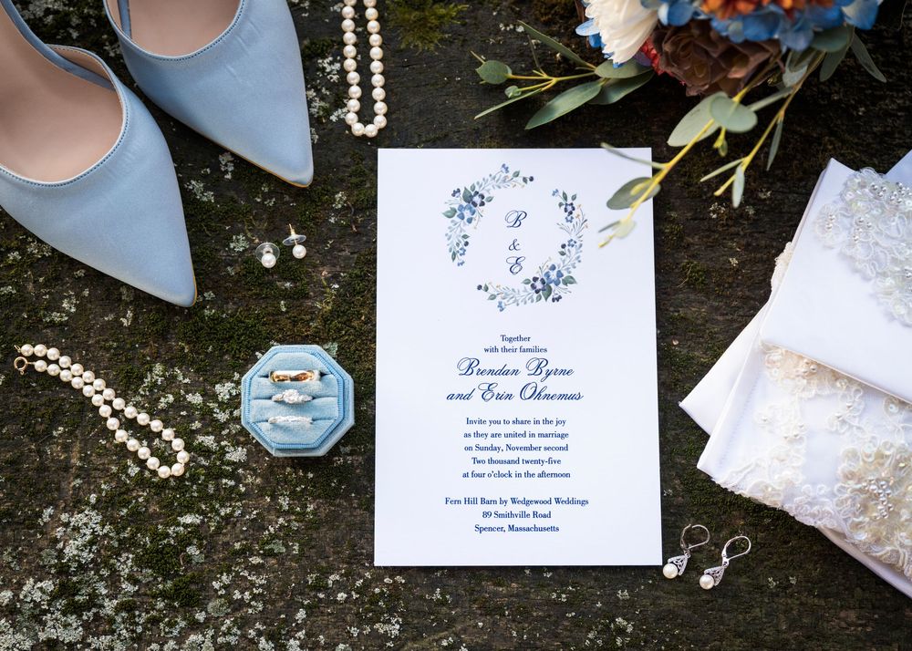 Wedding details flatlay with blue shoes, pearls, invitation and lace on dark surface.