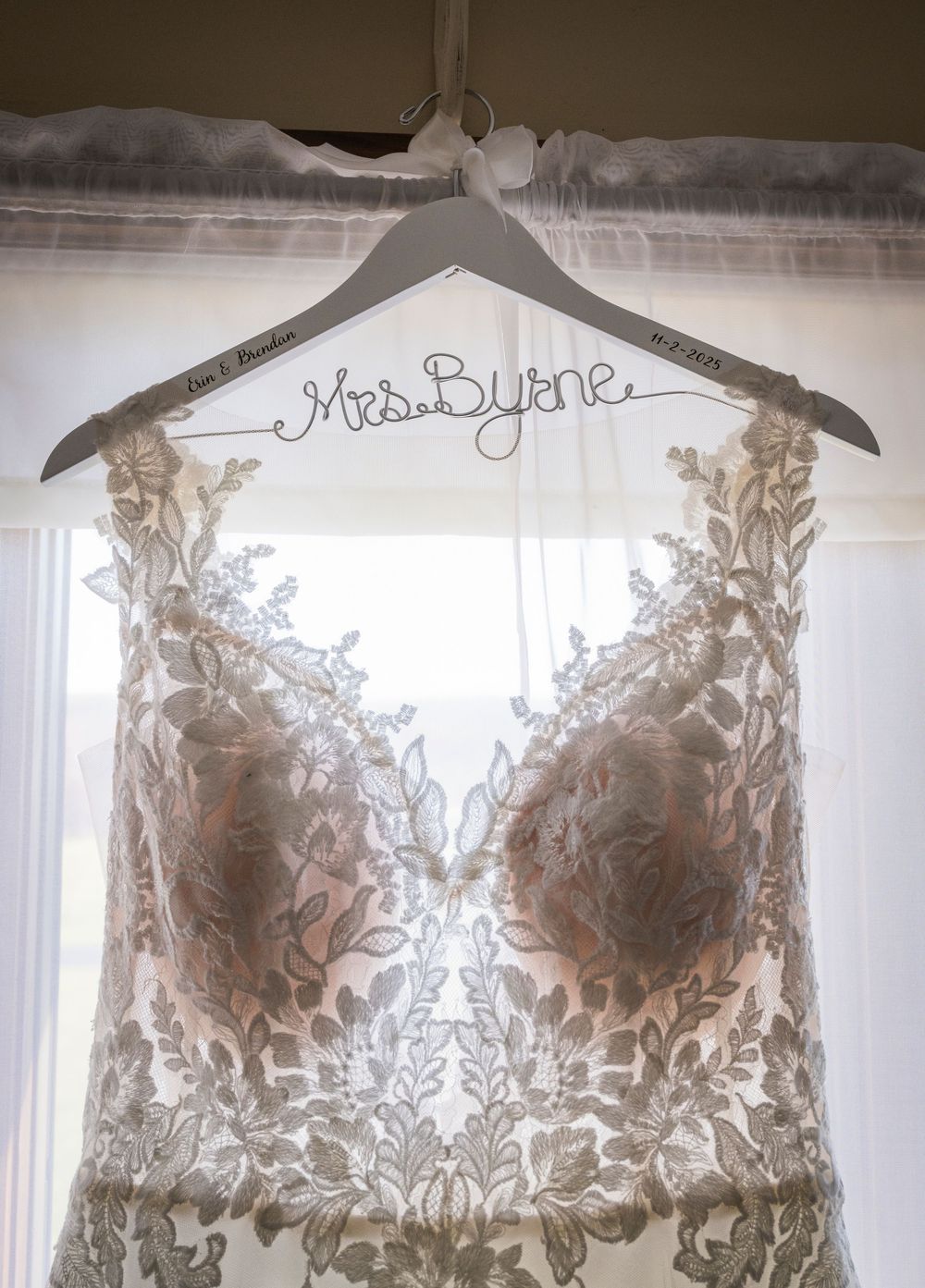 A detailed wedding dress with lace bodice hanging on a personalized hanger against a window.