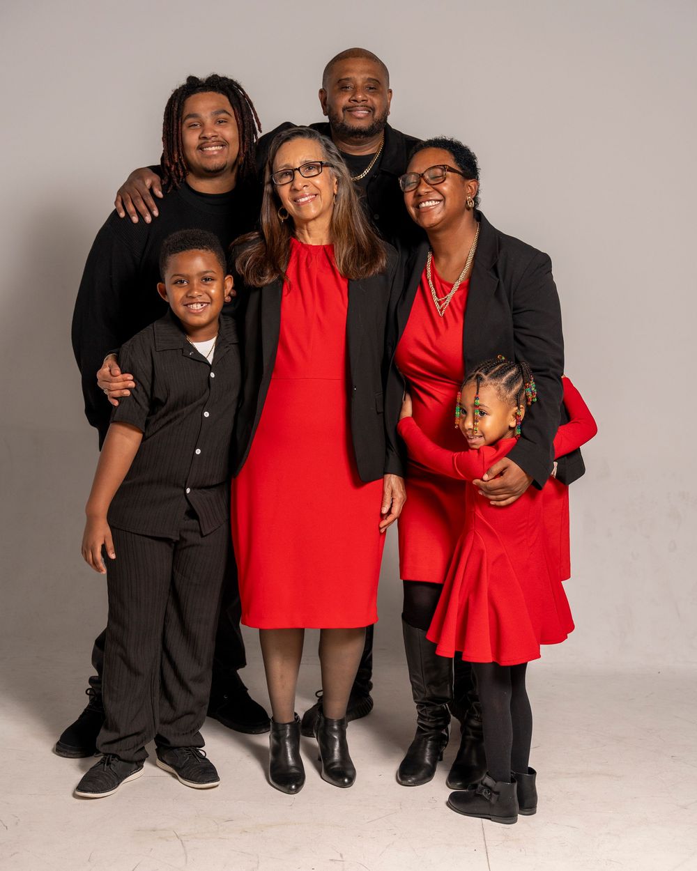 A cheerful family wearing coordinating red and black outfits poses together in a studio against a light gray background.