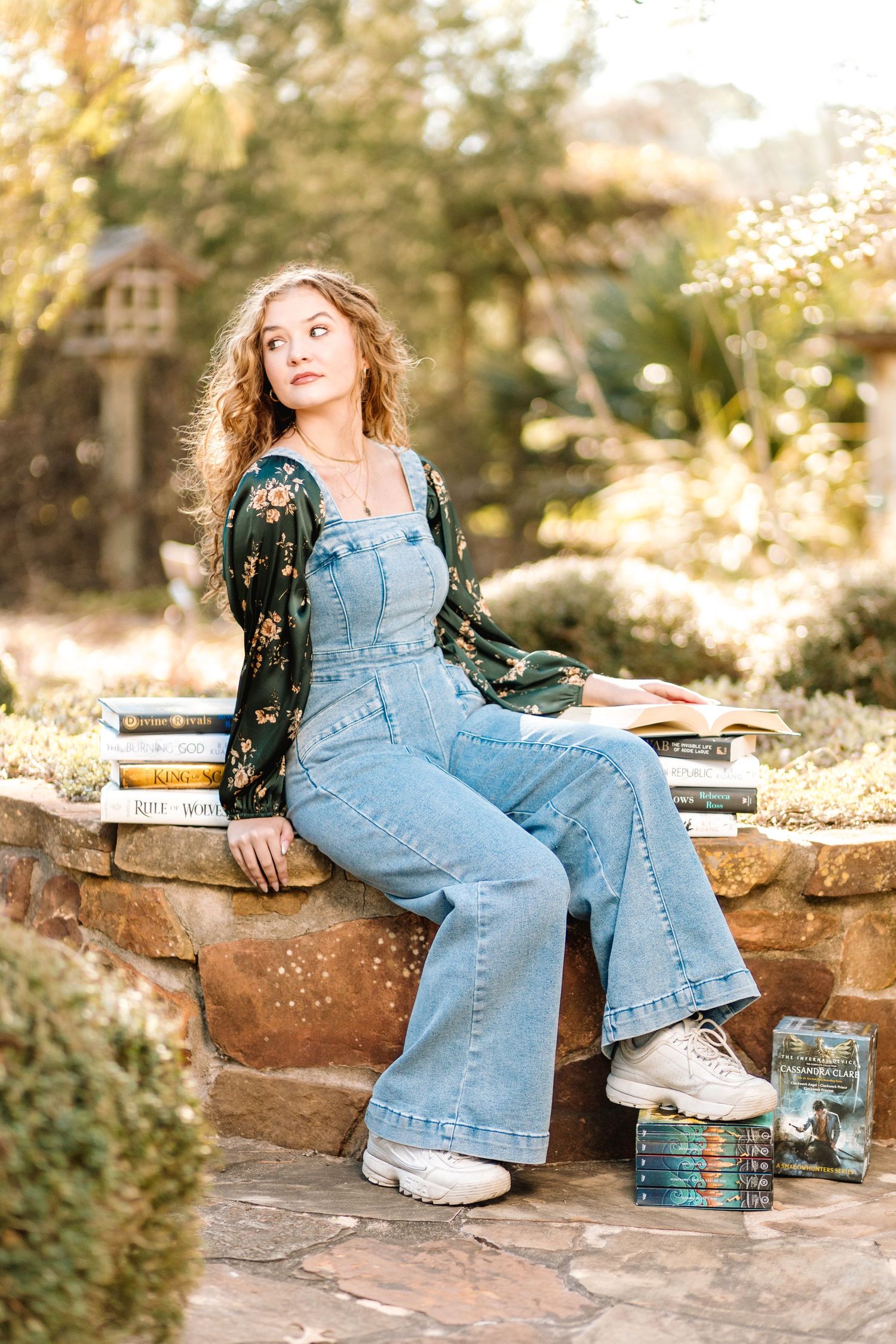 A person in denim overalls and floral blouse sits on stone steps in a sunlit garden setting with vintage decor.