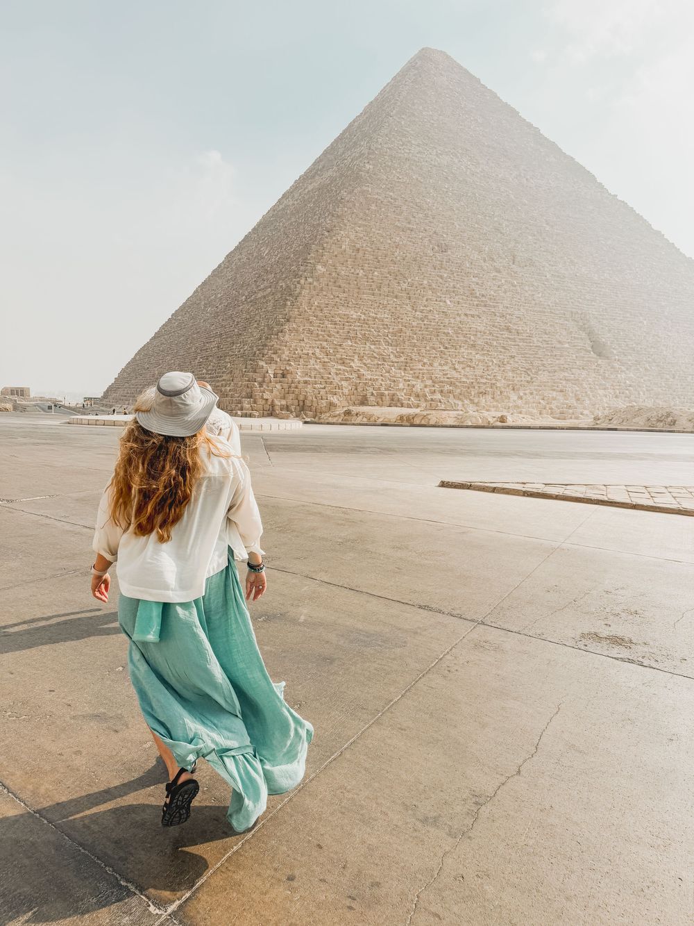 Tourist in flowy mint dress and white sweater walks near the Great Pyramid of Giza in Egypt.