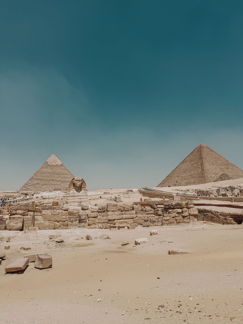 Ancient Egyptian pyramids and Sphinx rise from sandy ruins under teal sky at the historic Giza pyramid complex.