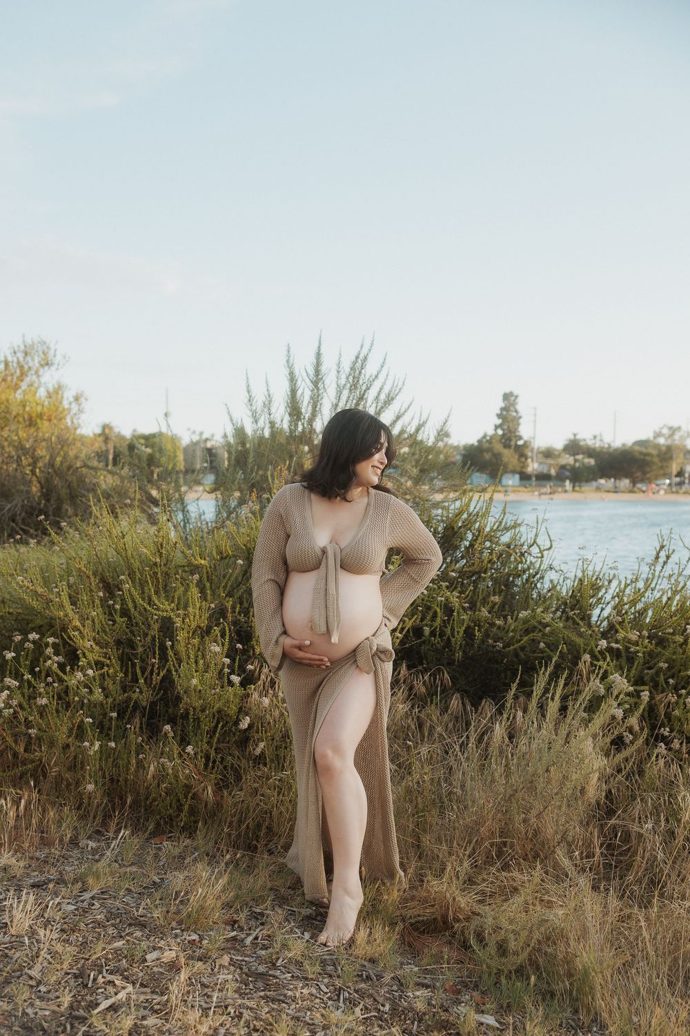 Scenic outdoor maternity photoshoot at sunset near a waterfront with natural grasses and warm lighting.