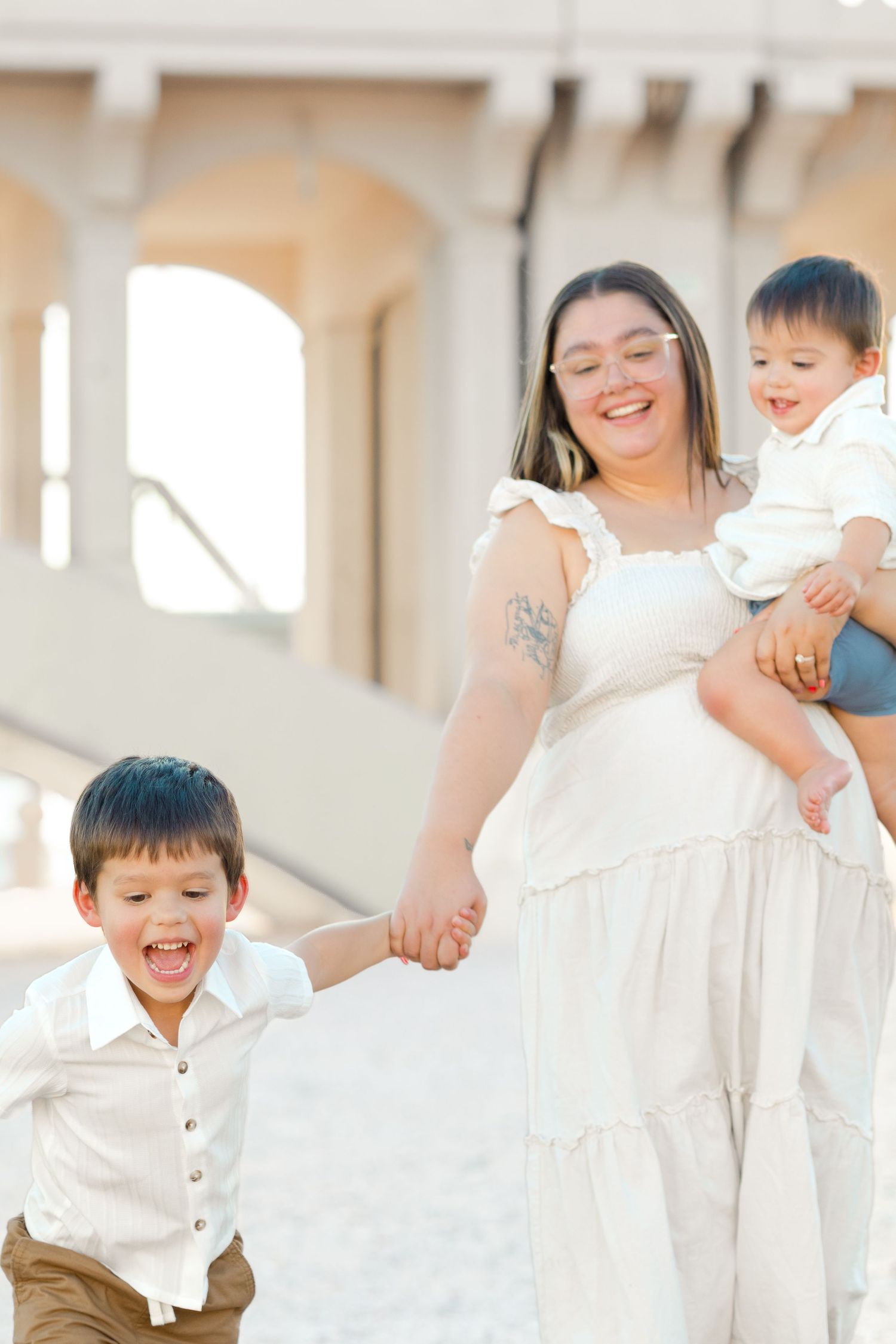 Light and airy family photo take by Arizona family photographer with mom holding infant son and toddler son holding hands walking with mom.