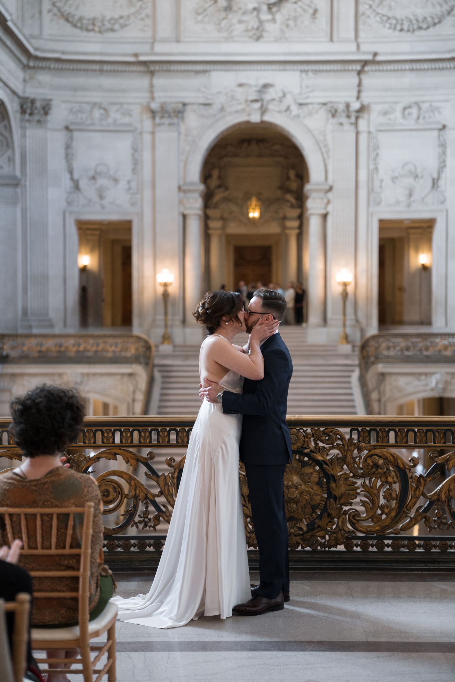 A couple shares a romantic moment in front of an elegant marble staircase in a grand historic building.