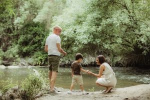 Family of three enjoying a peaceful moment by a forest creek, with parents and young child playing near the water.