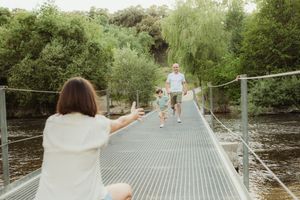 Family walking on a suspended footbridge over a river surrounded by lush green trees on a sunny day.