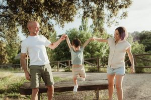 Family swinging young child outdoors in sunny park, parents holding hands with toddler between them.