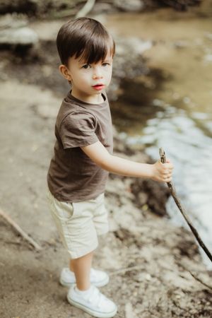 Young toddler boy in brown shirt and khaki shorts holding a stick near a stream outdoors.