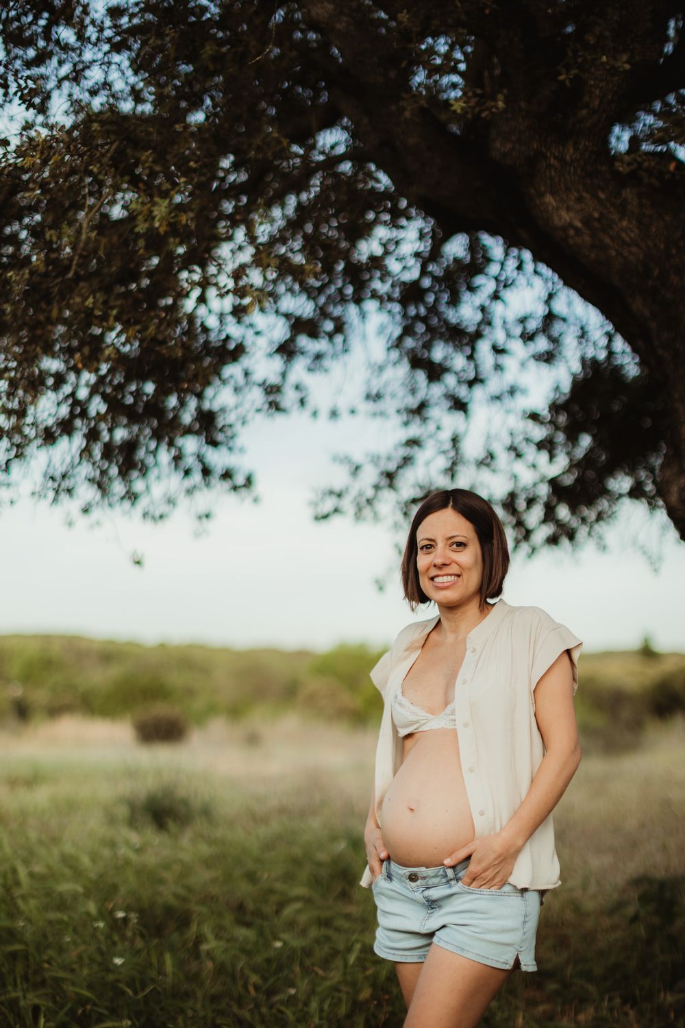 Smiling pregnant woman in denim shorts and open white top cradles her baby bump outdoors under a tree.