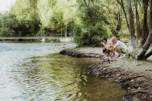 Father and child crouching by a peaceful river bank surrounded by lush green trees in a park.