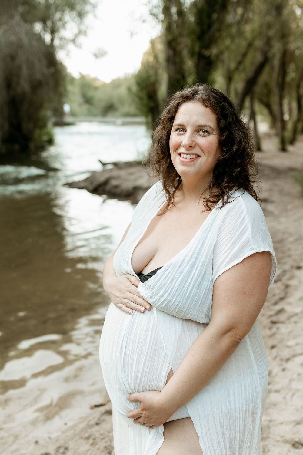Maternity portrait of an expectant mother in white dress by a river.
