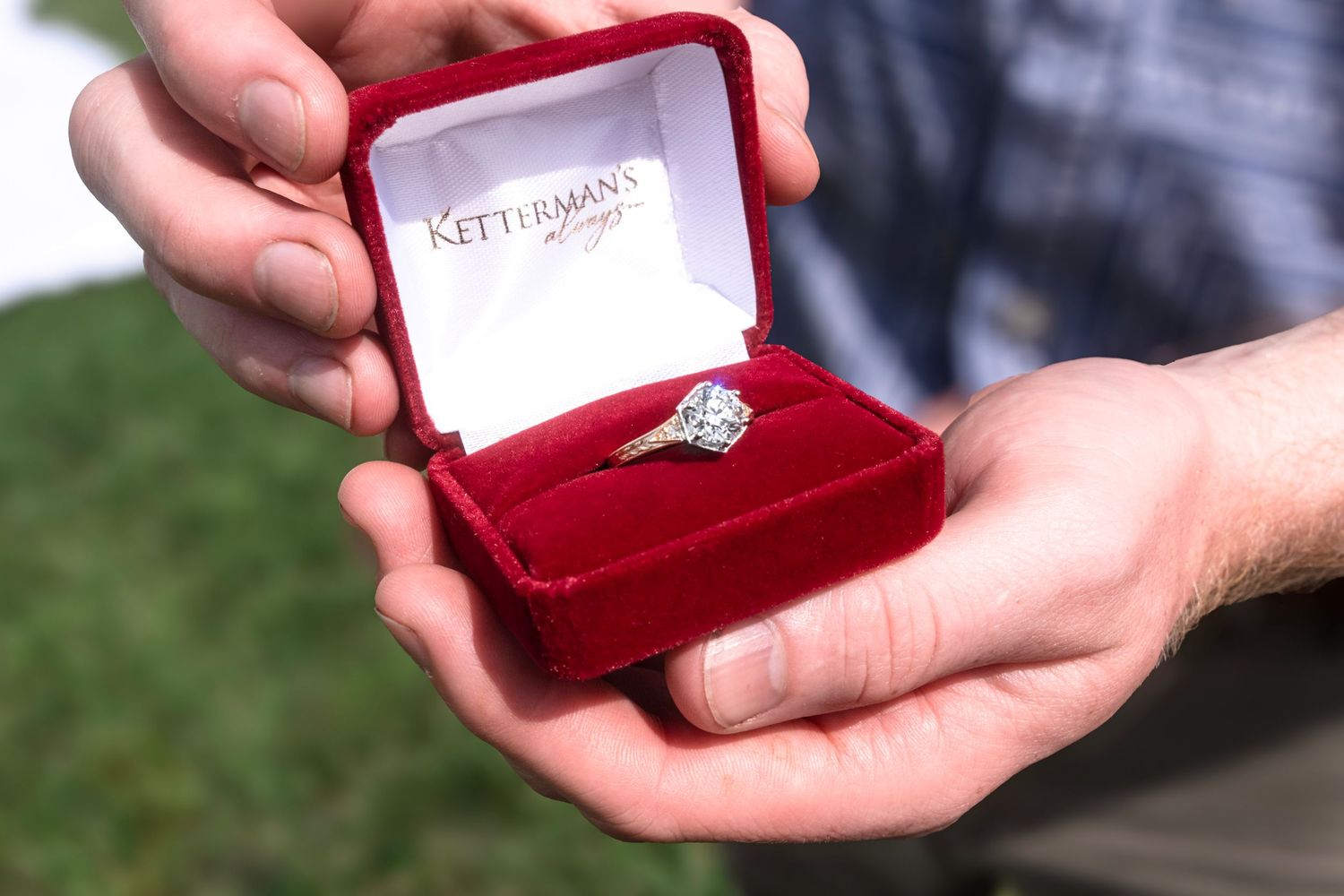 Close-up view of hands holding a red velvet ring box as it's opened to reveal an engagement ring.